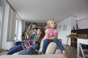 Porträt eines kleinen Mädchens, das auf der Rückenlehne einer Couch sitzt, mit ihrer Familie im Hintergrund - RBF003421