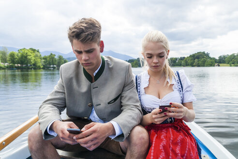 Deutschland, Bayern, Porträt eines jungen Paares, das in einem Ruderboot auf dem Staffelsee sitzt und auf seine Handys schaut - TCF004867