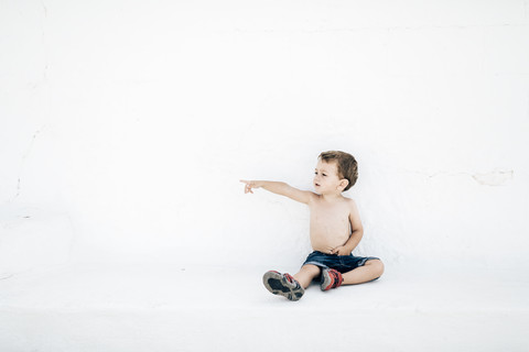 Spanien, Menorca, kleiner Junge ohne Hemd, der vor einer weißen Wand sitzt und auf etwas in der Ferne zeigt, lizenzfreies Stockfoto