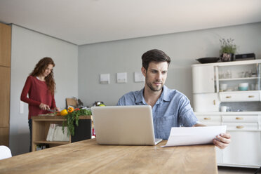 Mann zu Hause mit Laptop und Frau im Hintergrund, die Gemüse schneidet - RBF003546