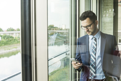 Junger Geschäftsmann am Fenster, der auf sein Handy schaut - UUF005409