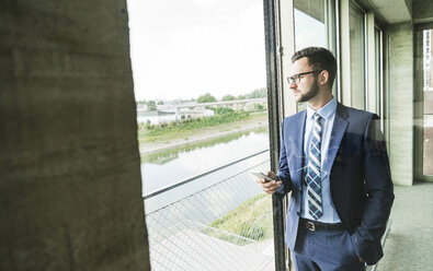 Junger Geschäftsmann mit Handy schaut aus dem Fenster - UUF005411