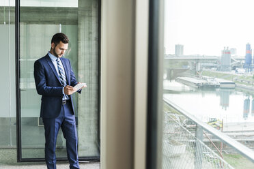 Young businessman at the window looking on digital tablet - UUF005414