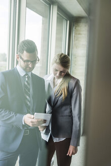 Young businessman and businesswoman at the window looking on digital tablet - UUF005416