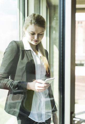 Lächelnde junge Geschäftsfrau am Fenster, die auf ihr Mobiltelefon schaut, lizenzfreies Stockfoto