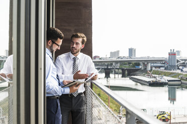 Two young businessmen with documents and digital tablet discussing on balcony - UUF005468