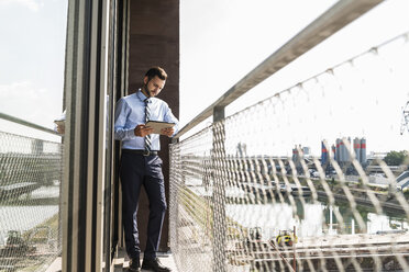 Junger Geschäftsmann steht auf einem Balkon und schaut auf ein digitales Tablet - UUF005470
