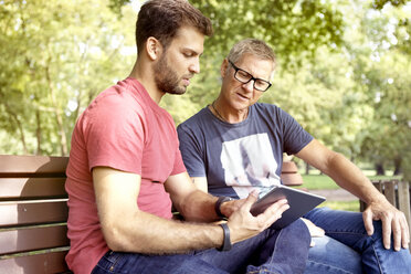 Zwei Männer sitzen auf einer Parkbank und schauen auf ein digitales Tablet - SEGF000414