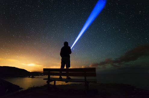 Spanien, Ortigueira, Loiba, Silhouette eines auf einer Bank stehenden Mannes unter Sternenhimmel mit blauem Strahl - RAEF000391