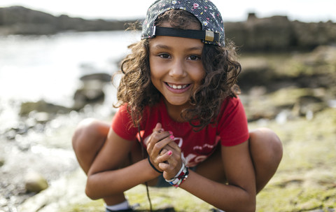 Spain, Gijon, portrait of smiling little girl crouching at rocky coast stock photo