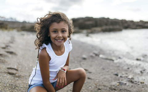 Spain, Gijon, portrait of smiling little girl crouching on the beach stock photo