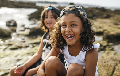 Spanien, Gijon, Porträt eines lachenden kleinen Mädchens und ihrer Freundin im Hintergrund, die an einer Felsenküste sitzen - MGOF000543