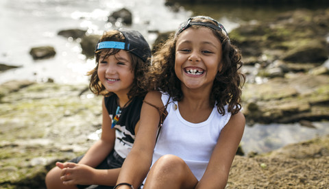 Spain, Gijon, portrait of smiling little girl and her friend in the background sitting at rocky coast stock photo