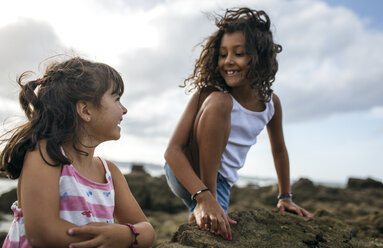 Spain, Gijon, portrait of two smiling little girls playing at rocky coast - MGOF000549