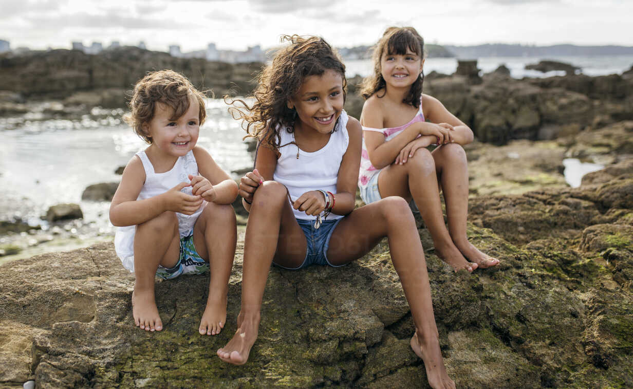Spain, Gijon, group picture of three little girls sitting at rocky coast  stock photo