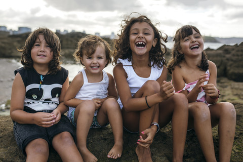 Spanien, Gijon, Gruppenbild von vier kleinen Kindern, die an der felsigen Küste sitzen und Spaß haben, lizenzfreies Stockfoto
