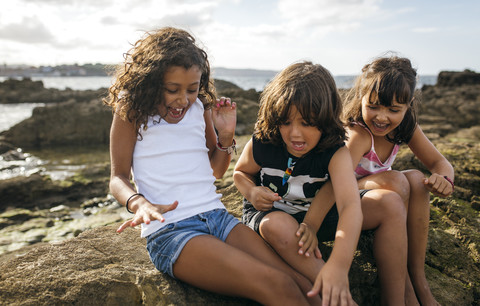 Spain, Gijon, group picture of three excited little children sitting at rocky coast stock photo