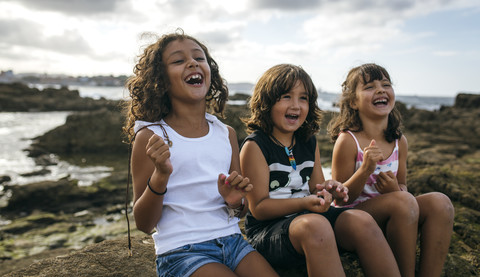 Spain, Gijon, group picture of three laughing little children sitting at rocky coast stock photo