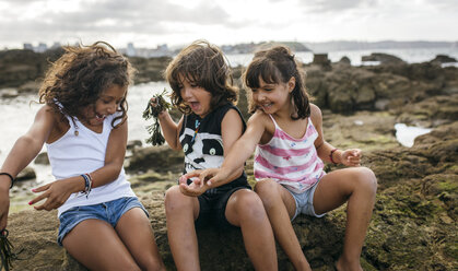 Spain, Gijon, group picture of three excited little children sitting at rocky coast - MGOF000564