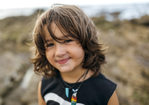 Spanien, Gijon, Porträt eines lächelnden kleinen Jungen mit braunem Haar an der felsigen Küste, lizenzfreies Stockfoto