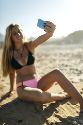 Young woman sitting on the beach taking a selfie with her smartphone - MGOF000571