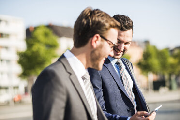 Two young businessmen walking in city, looking at mobile phone - UUF005559