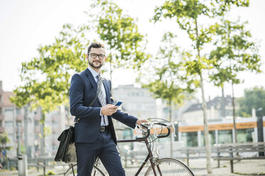 Junger Geschäftsmann, der ein Fahrrad schiebt und ein Smartphone in der Hand hält - UUF005563