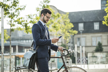 Young businessman pushing bicycle, holding smart phone - UUF005564