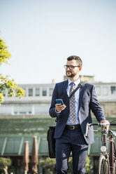 Young businessman pushing bicycle, holding smart phone - UUF005565