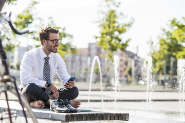 Young businessman sitting cross-legged on bench, using smartphone - UUF005596