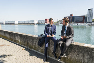 Two young businessmen sitting on wall by river, working - UUF005618