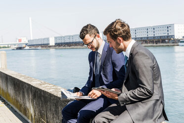 Two young businessmen sitting on wall by river, working - UUF005632
