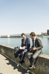 Two young businessmen sitting on wall by river, working - UUF005620