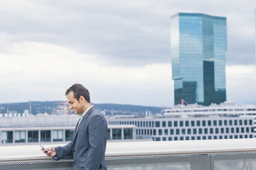 Businessman on urban balcony text messaging on cell phone - BZF000217