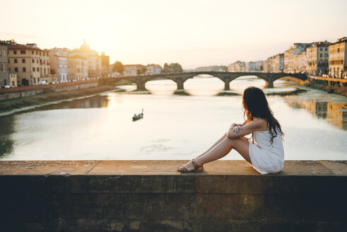 Italy, Florence, woman wearing white summer dress sitting on a bridge at sunset - GEMF000324