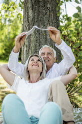 Elderly couple sitting at tree reading book - RKNF000196