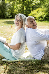 Smiling elderly couple sitting back to back in grass - RKNF000201