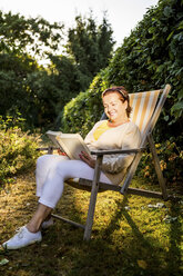Lächelnde reife Frau, die im Liegestuhl im Garten ein Buch liest - RKNF000217