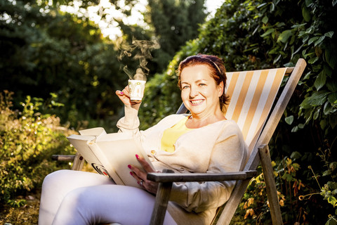 Lächelnde reife Frau liest ein Buch und trinkt Kaffee im Liegestuhl im Garten, lizenzfreies Stockfoto