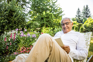 Smiling senior man reading book in garden - RKNF000238
