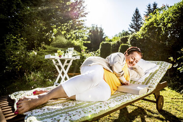 Lächelnde reife Frau beim Lesen eines Buches in der Gartenlounge - RKNF000256