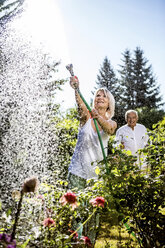 Lächelnde reife Frau gießt Blumen im Garten mit Mann im Hintergrund - RKNF000292