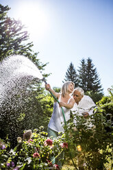 Smiling mature woman watering flowers in garden with man kissing her shoulder - RKNF000293