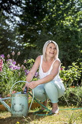 Smiling mature woman with hose and watering can in garden - RKNF000295
