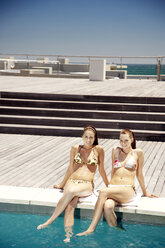 Zwei glückliche junge Frauen sitzen am Pool - TOYF001386