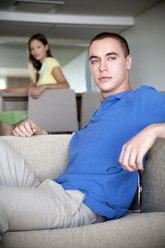 Junger Mann sitzt auf einer Couch mit einer Frau im Hintergrund - TOYF001388