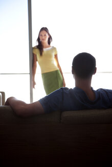 Junger Mann auf der Couch mit Blick auf seine Freundin am Fenster - TOYF001353
