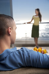 Junger Mann auf Couch mit Freundin auf Balkon am Meer stehend - TOYF001354
