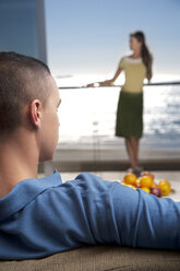 Young man on couch with girlfriend standing on balcony by the sea - TOYF001354