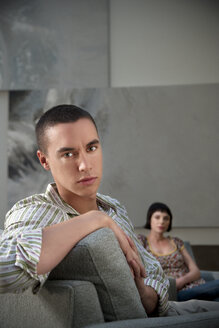 Junger Mann sitzt auf einer Couch mit einer Frau im Hintergrund - TOYF001360